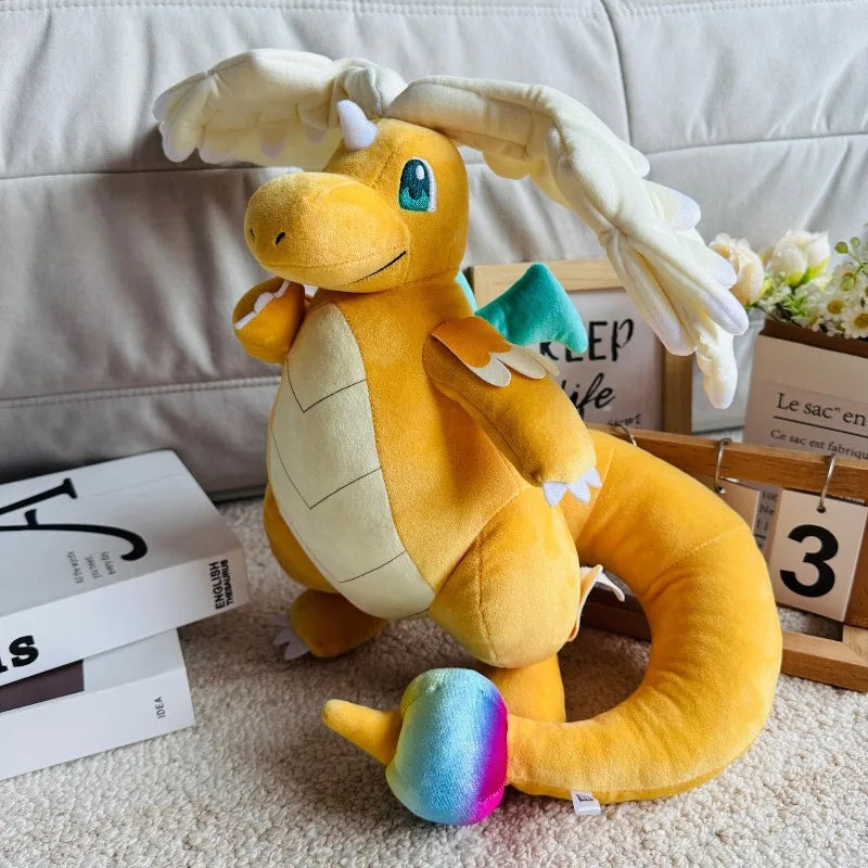 Pokemon Dragonite Plush Toy – Large Soft Stuffed Doll A yellow and cream dragon plush toy with a multicolored rainbow tail, posed on a beige carpet in a styled room setting with books, a calendar, and a sofa in the background.
