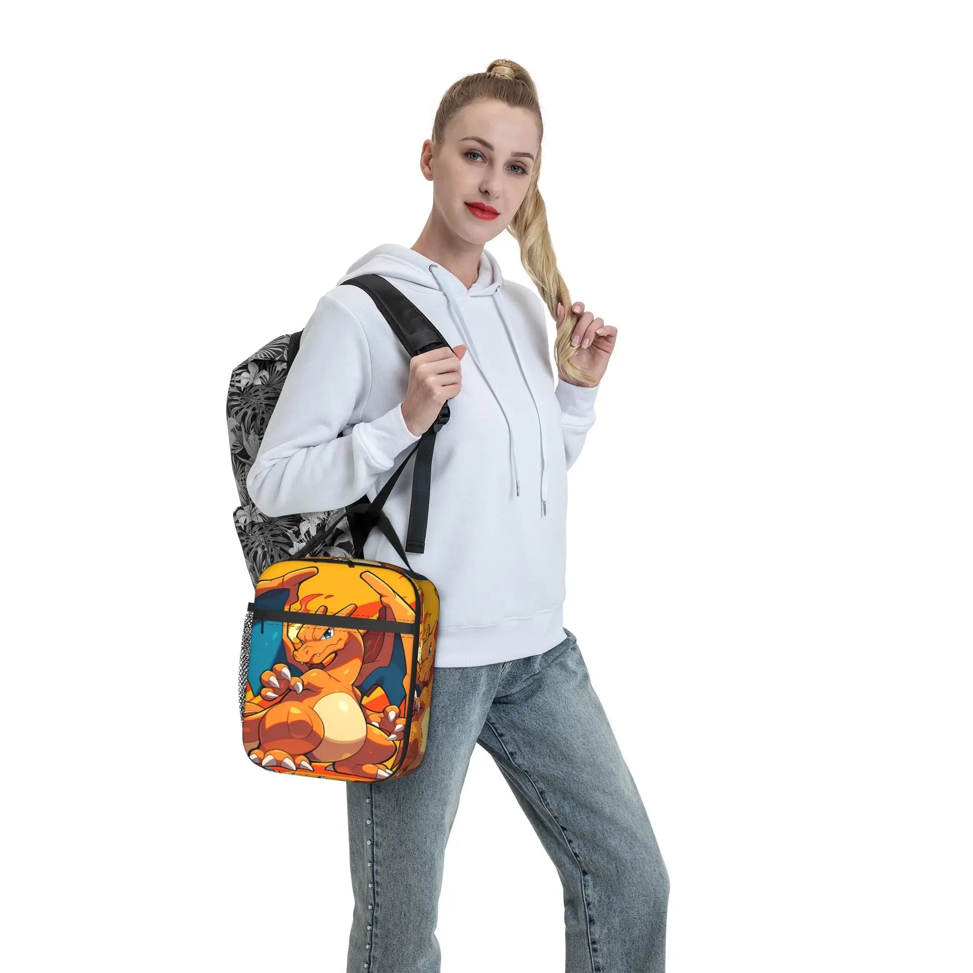 Pokemon Charizard Fire Burst Lunch Bag – Waterproof Insulated Tote A woman wearing a white hoodie and blue jeans models a small rectangular crossbody bag with a colorful cartoon-style print. The bag features orange, yellow, and blue graphics with black trim and a black-and-white patterned secondary bag partially visible behind it, set against a plain white background.