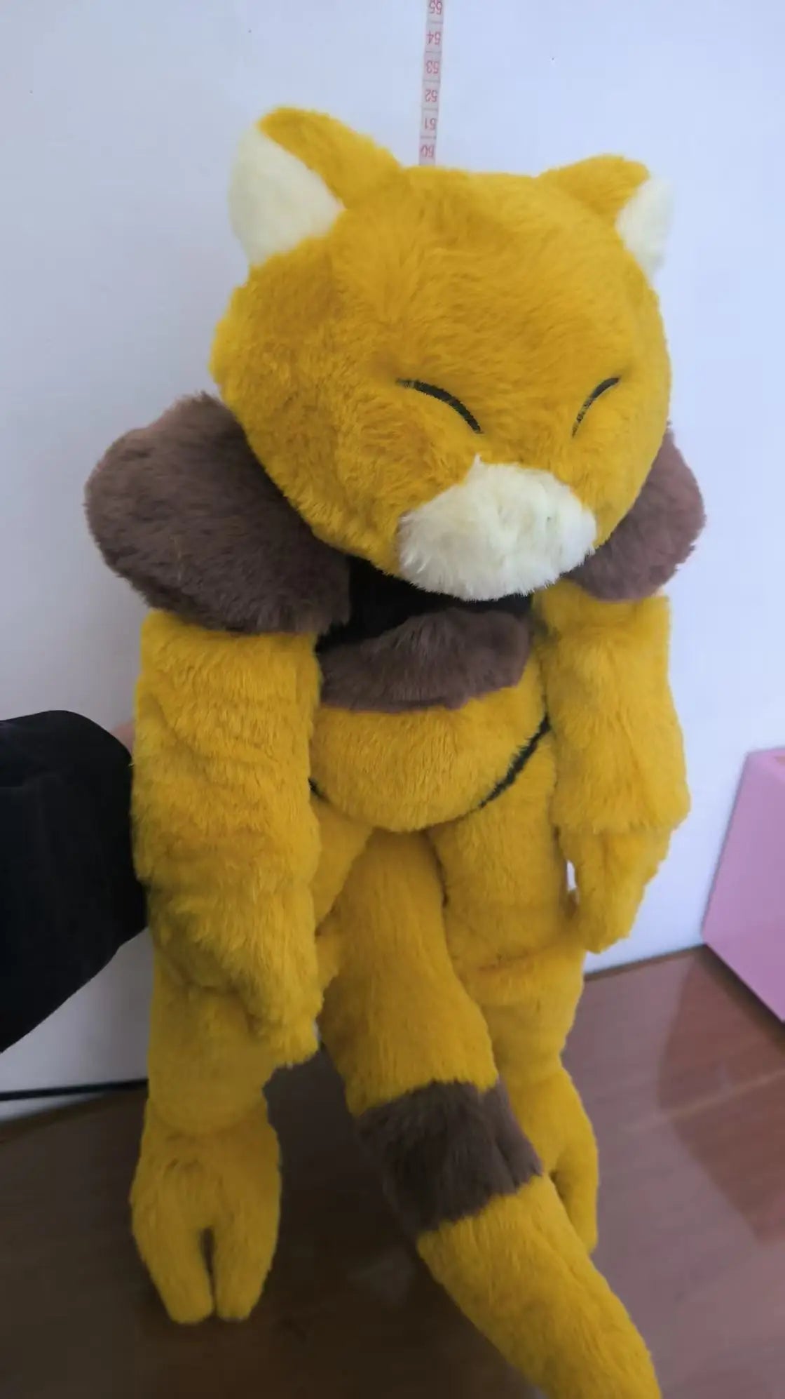 Pokemon Abra Plush Toy – Soft Warm Companion Stuffed Doll A yellow plush toy with brown and cream accents, resembling a stylized animal character, standing on a wooden floor against a plain wall.
