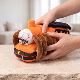A plush toy shaped like a spider-themed pumpkin in orange, black, and brown, being held and squeezed by two hands on a stone tabletop with a blurred indoor background.