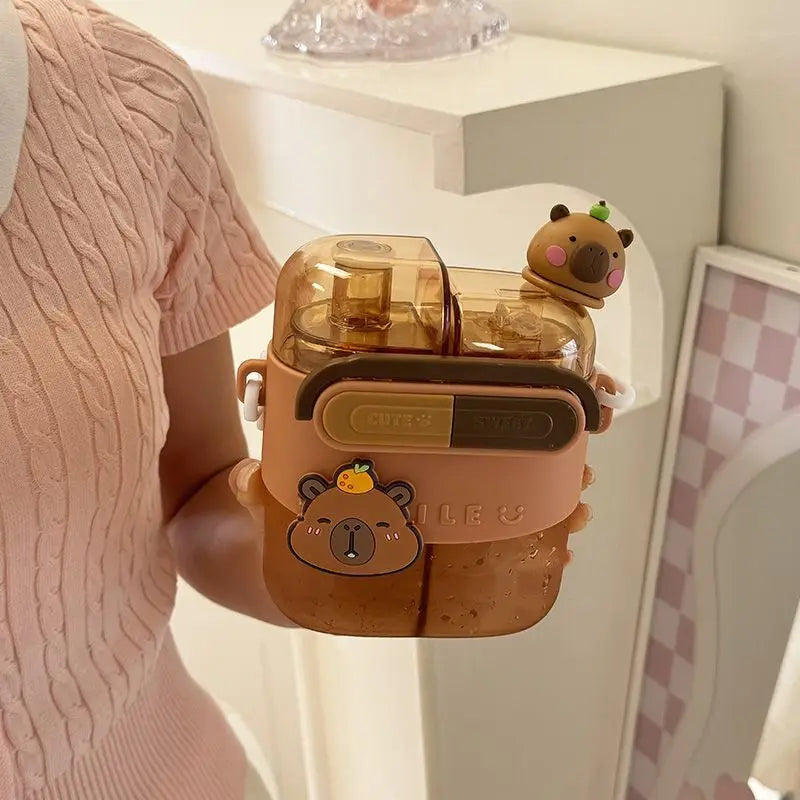 Cute Capybara Kids Water Bottle – Large Capacity Straw Cup A person in a pink ribbed top holds a double-straw water bottle with a peach-colored silicone sleeve, cartoon bear graphics, and a small capybara figure on top, set against a white shelf and pink-and-white checkered mirror.