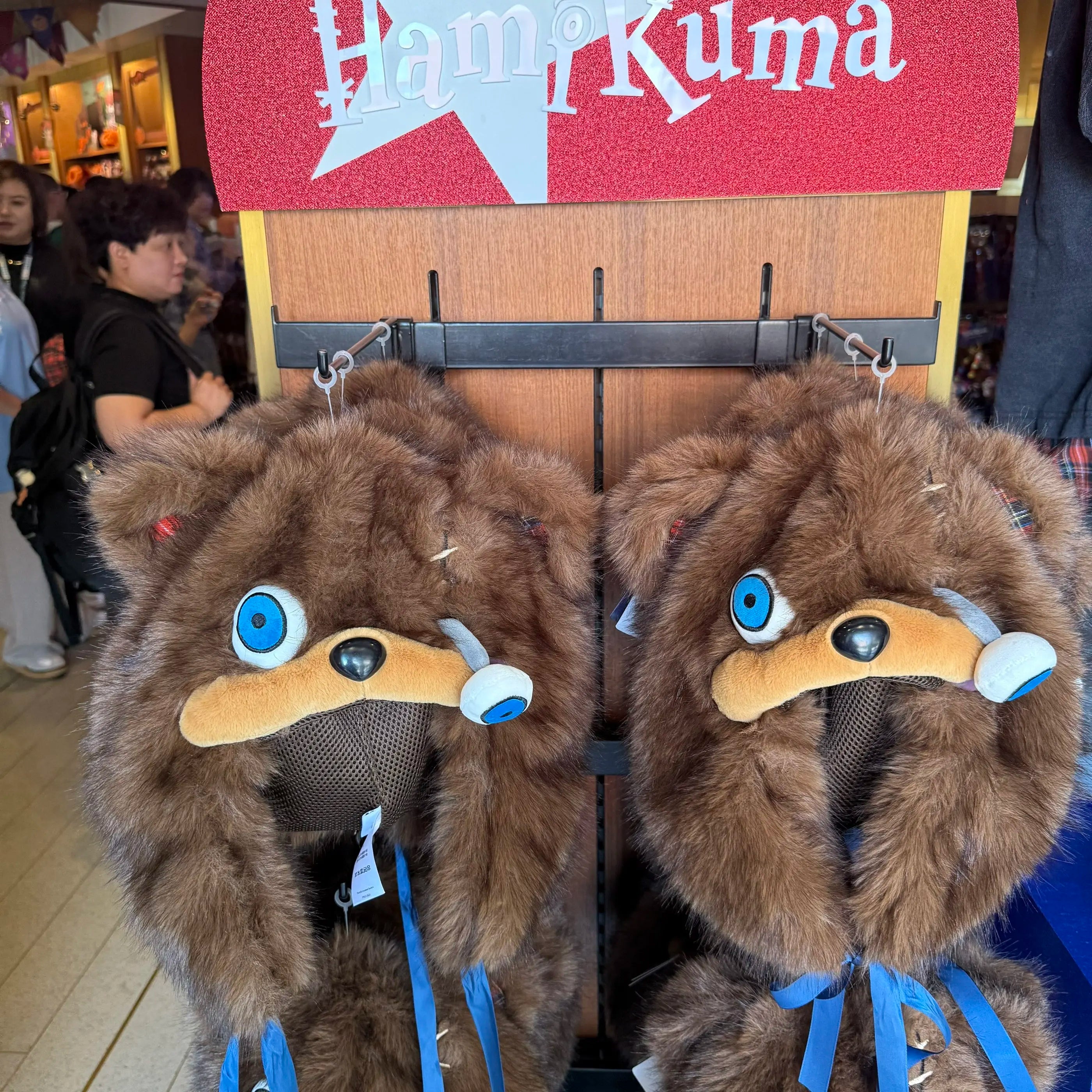 Hamikuma Bear Plush Hooded Halloween Trapper Hat Two brown furry character hats with blue eye patches and blue ribbons hanging on a display stand in a store. A red sign reading 'Ham Kuma' is visible above, with people blurred in the background.