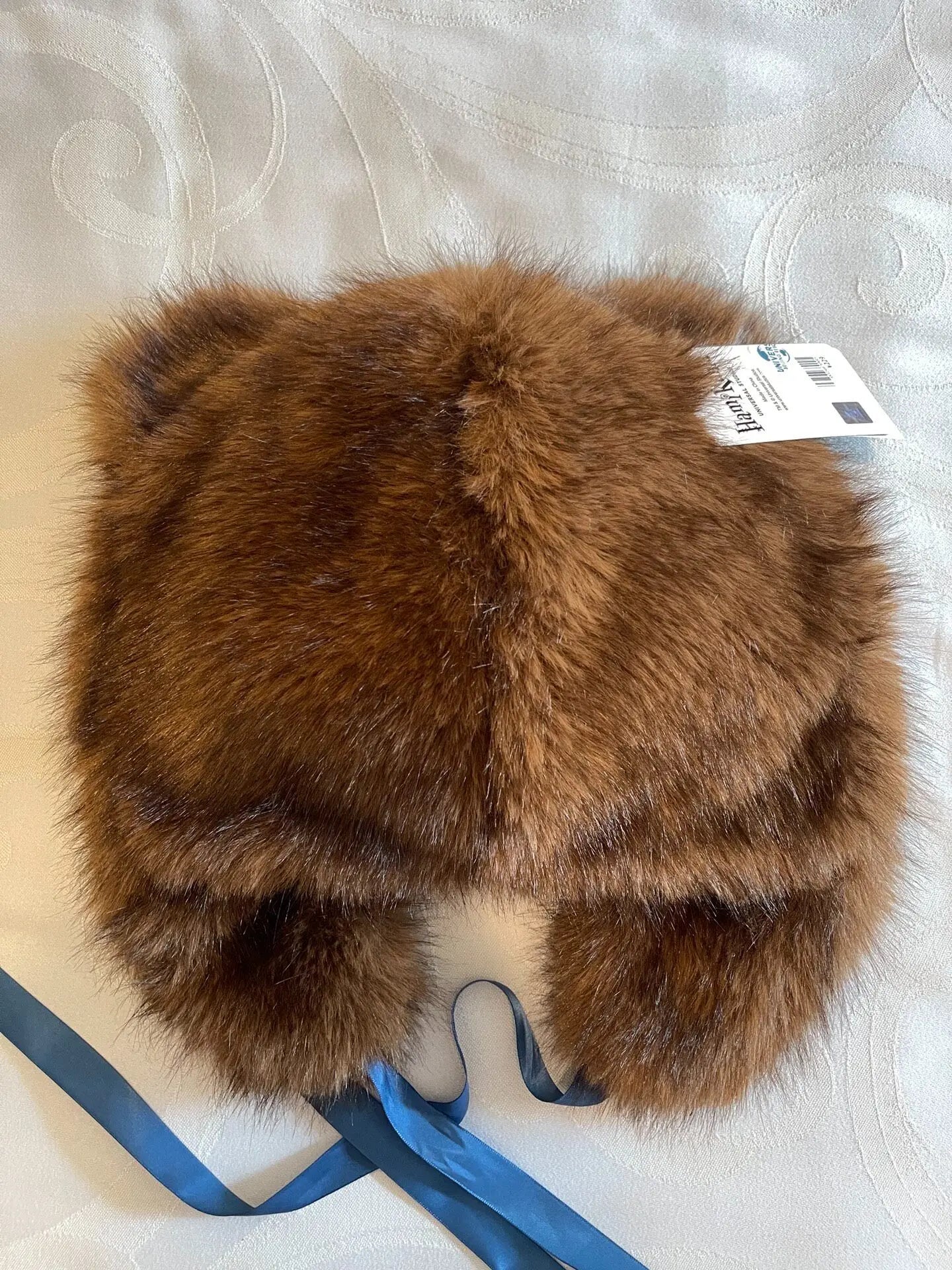 Hamikuma Bear Plush Hooded Halloween Trapper Hat A brown faux fur trapper hat with ear flaps and blue ribbons, shown from the back on a white patterned fabric background.