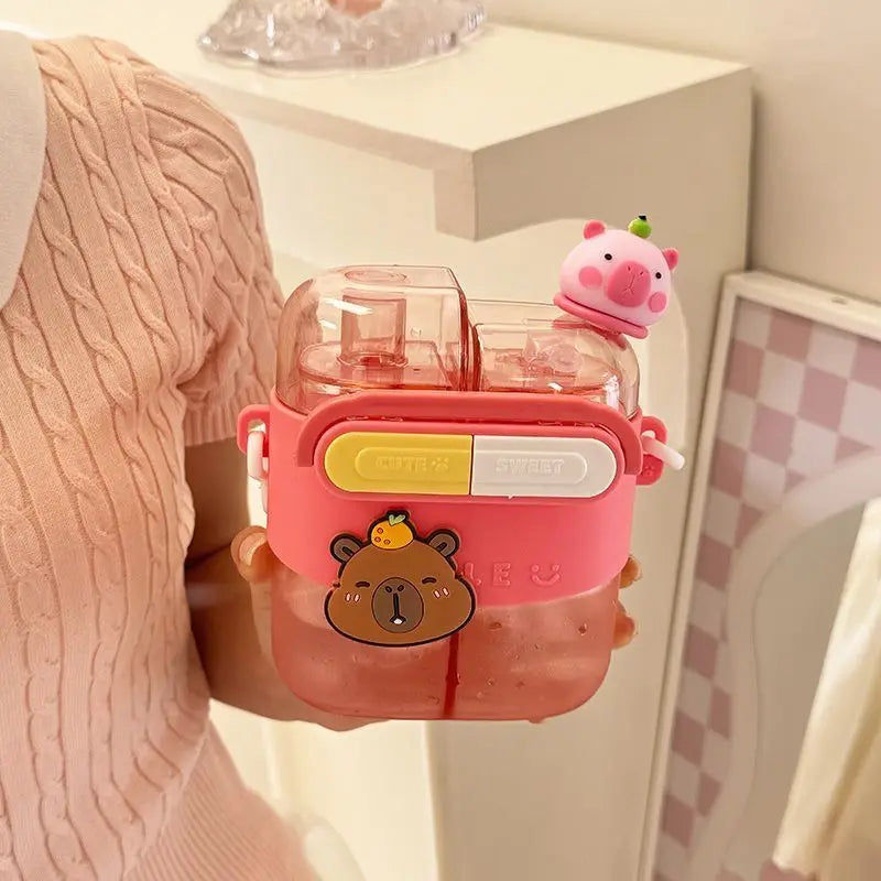 Cute Capybara Kids Water Bottle – Large Capacity Straw Cup A person in a pink cable-knit sweater holds a pink water bottle with a transparent cap. The bottle has a pink silicone sleeve with a cartoon bear face, a yellow-and-white flip tab, and a small pink pig character on top, shown in a bright indoor setting.