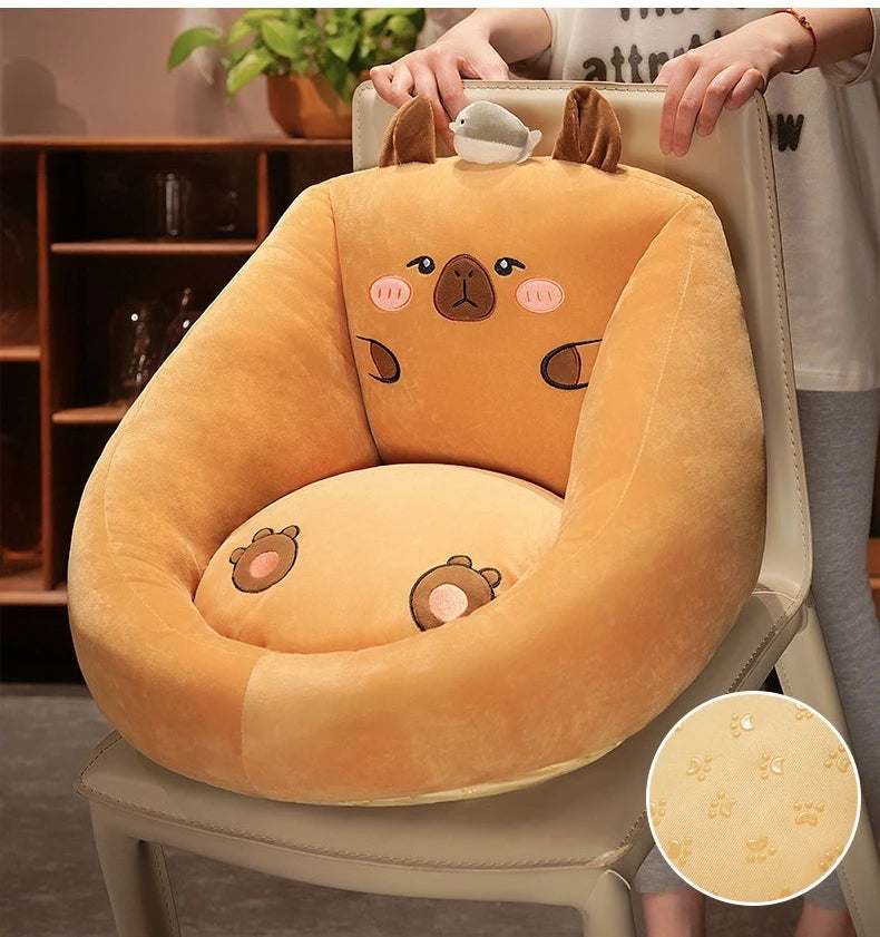Capybara Floor Cushion – Futon Seat for Living Room & Bedroom A plush capybara-shaped baby support cushion in warm tan and orange-brown, shown being held over a chair in a home setting. The cushion features embroidered cute capybara face details and paw prints on the fabric, with a circular inset showing a close-up of the textured fabric.