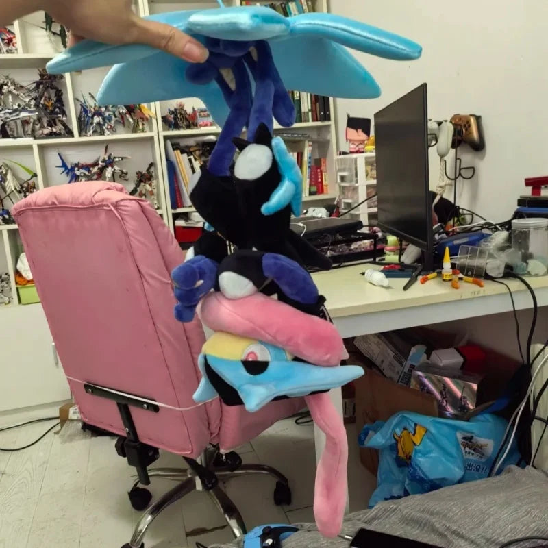 Pokemon Mega Plush Toy – Mega Lucario, Mega Greninja & Mega Starmie A person holds a stack of plush toys, including a blue winged character, a black cat-like plush, a pink bird-like plush, and another blue/yellow plush, in a cluttered home office or playroom with a pink chair, shelves, and computer desk visible.