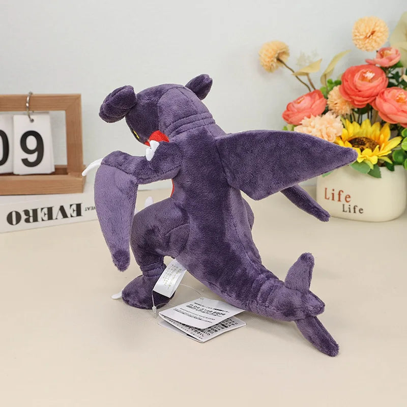 Pokemon Garchomp Plush Toy – Garchomp, Gabite & Gible Stuffed Animal Purple plush toy with a tag on a light surface with flowers in the background