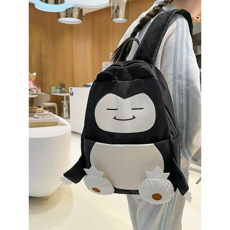 Snorlax Pokemon Backpack Large Capacity Casual Bag A person wearing a light gray and blue tracksuit carries a black and white plush penguin backpack in a modern indoor setting with warm wood walls and white furniture.
