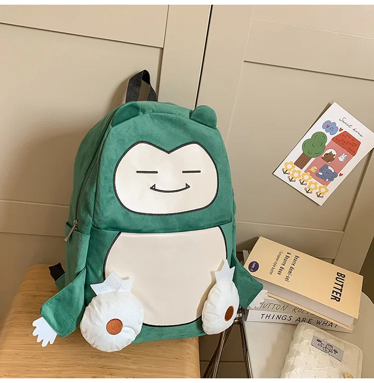 Snorlax Pokemon Backpack Large Capacity Casual Bag A green plush character backpack styled on a wooden table next to books and a card, featuring a white belly, white hands, and a smiling face.