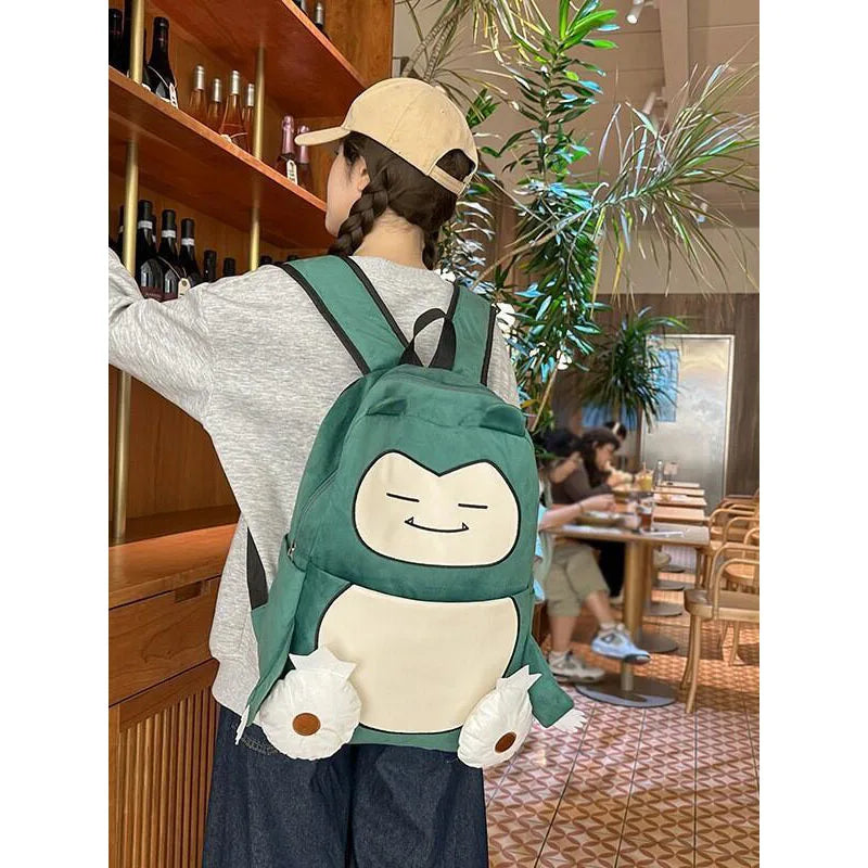 Snorlax Pokemon Backpack Large Capacity Casual Bag A person wearing a beige cap and gray sweatshirt is shown from behind in a restaurant, carrying a green and cream character backpack that resembles a cartoon creature with white flower-like accents.