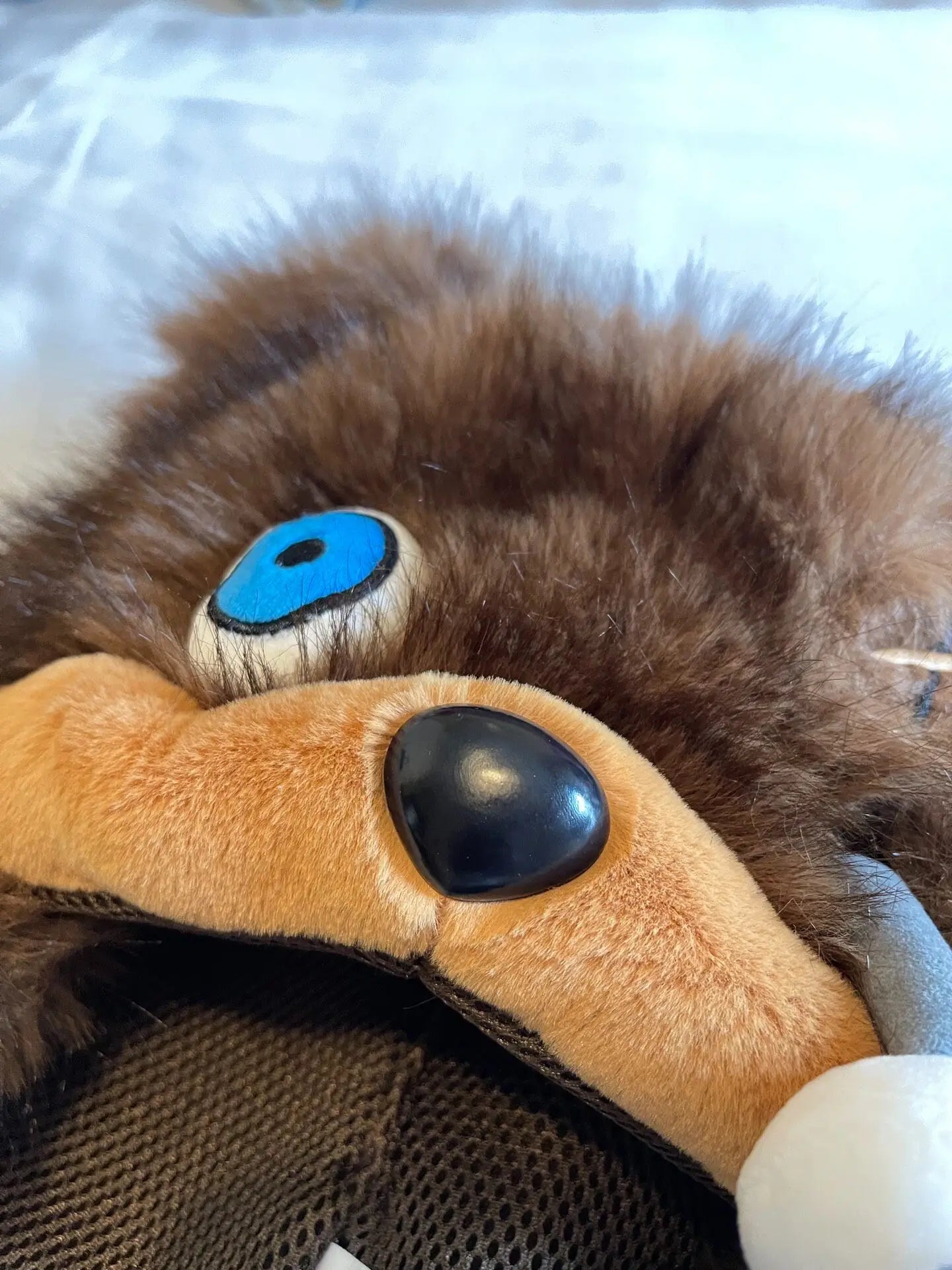 Hamikuma Bear Plush Hooded Halloween Trapper Hat Close-up of a plush toy or stuffed animal head with brown faux fur, tan fabric, a large black button eye, and a bright blue accent eye.