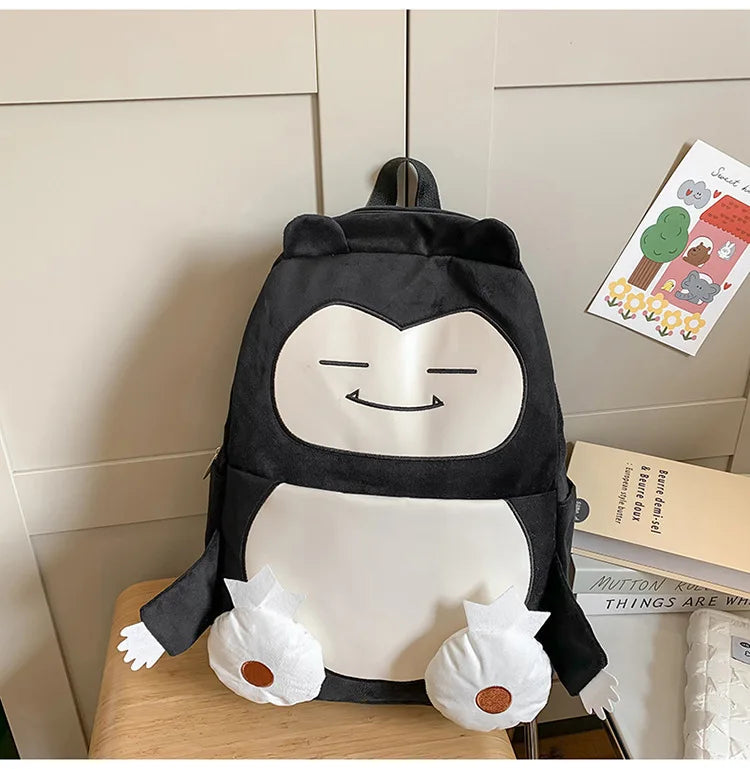 Snorlax Pokemon Backpack Large Capacity Casual Bag A black and white character backpack shaped like a sleeping creature with small white plush hands and feet, displayed on a wooden surface against a beige paneled wall.