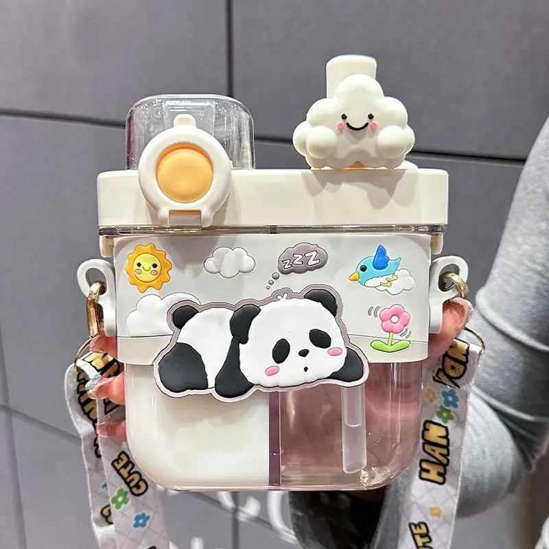 Capybara Kids Water Bottle – Cartoon Straw Cup A cute, transparent and white plastic water bottle with a carrying strap, decorated with a 3D panda, cloud, sun, and bird sticker. The bottle is held by a person wearing a gray long-sleeve shirt against a gray tiled wall background.