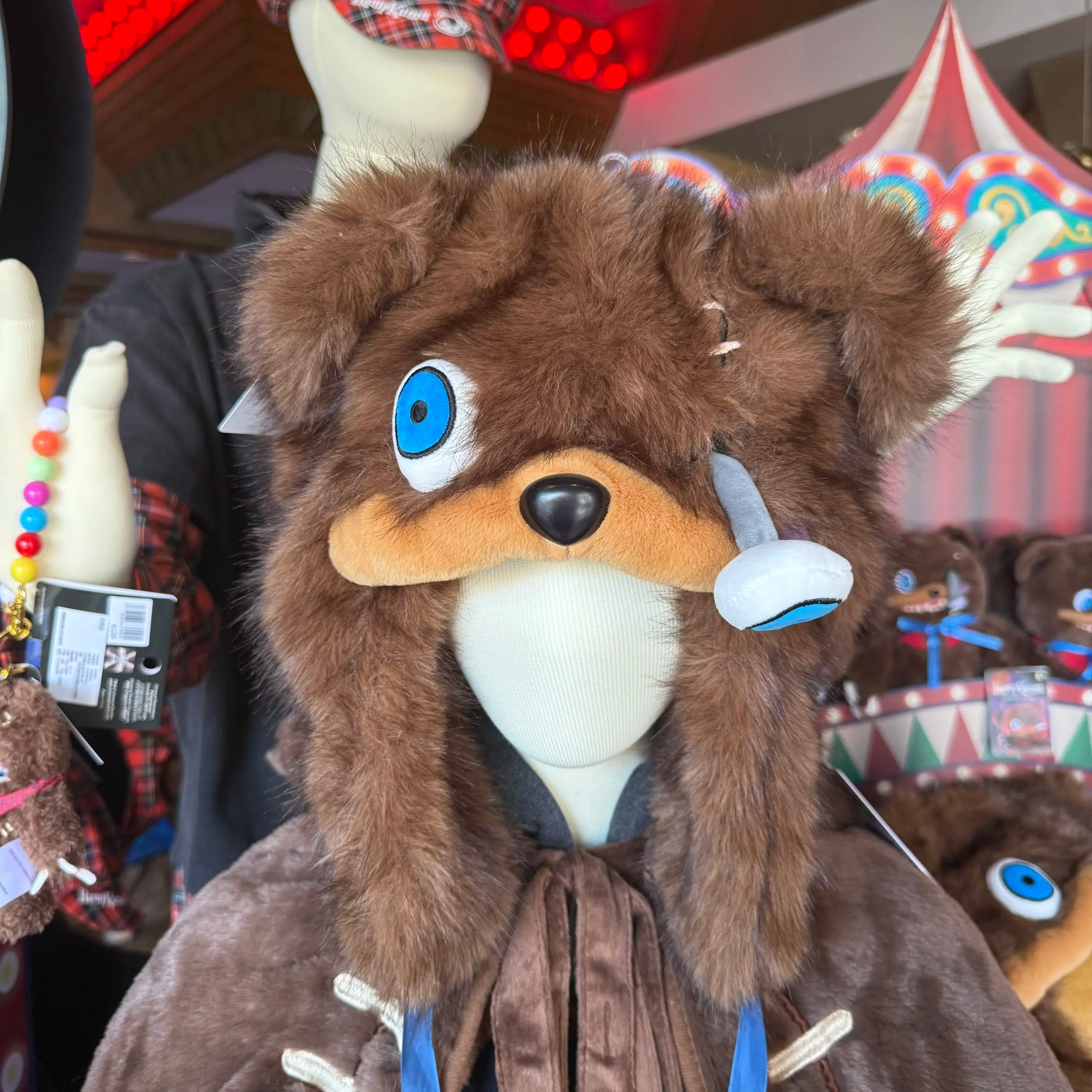 Hamikuma Bear Plush Hooded Halloween Trapper Hat A brown furry hooded costume or plush animal head with large blue eyes, a tan snout, and white accents, shown on a mannequin in a retail display with other toys and decorations in the background.