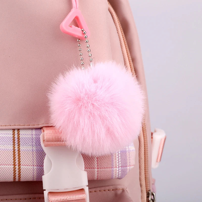 Capybara Bubble Tea Backpack – School Bag for Teens A pink fluffy pom-pom bag charm with a silver ball chain and pink plastic clip, attached to a light pink backpack with a plaid strap.