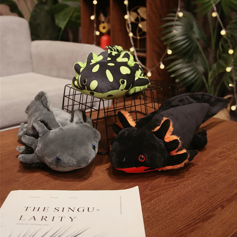 Cute Axolotl Plush Toy – Squishmallow Salamander Stuffed Animal Three plush axolotl toys arranged on a wooden table in a cozy indoor setting, with a gray plush in front, a black and neon green plush on a wire stand behind, and a black plush with orange accents on the right. A book and sofa are visible in the background.