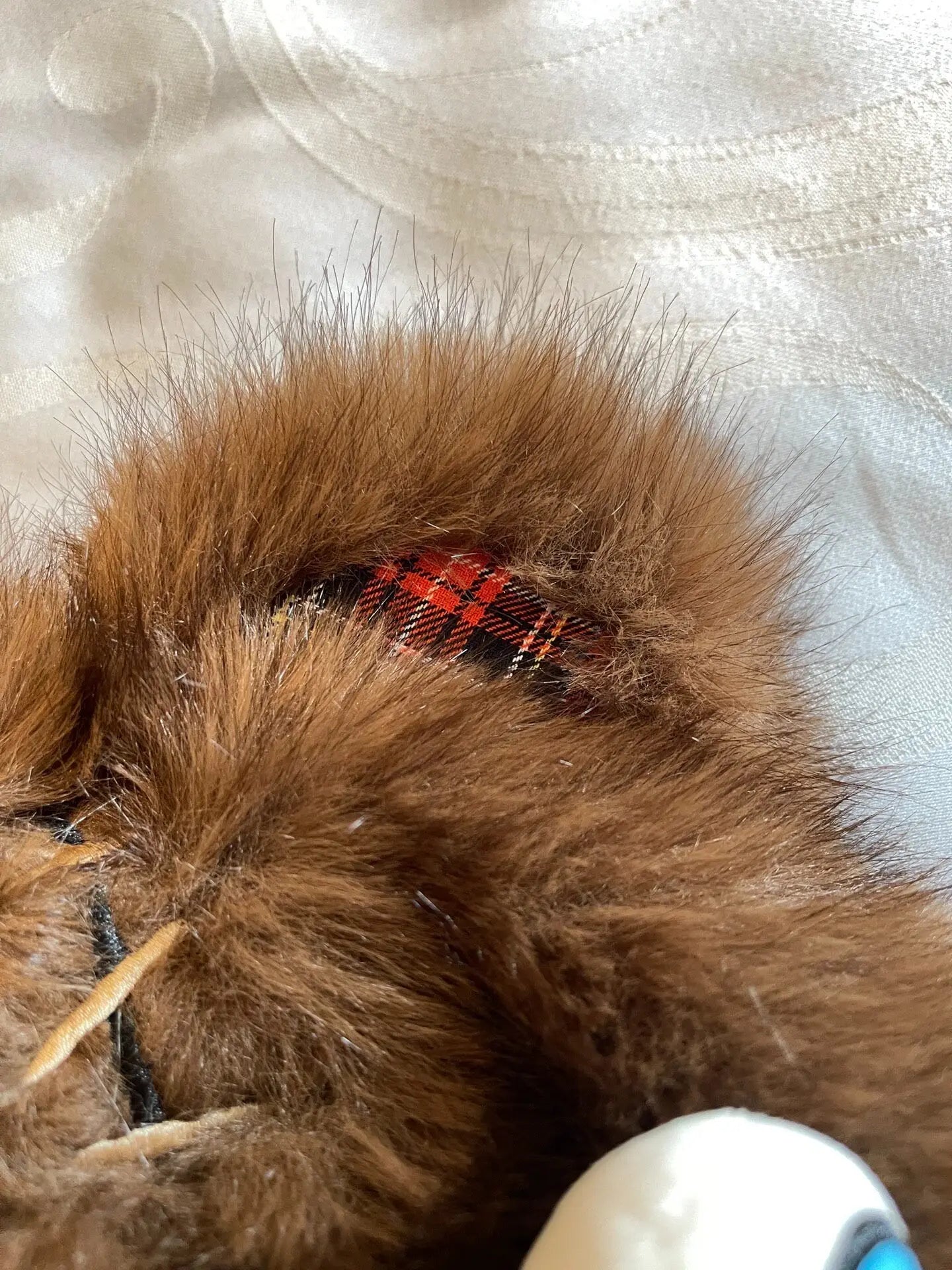 Hamikuma Bear Plush Hooded Halloween Trapper Hat A close-up of a brown faux fur accessory, likely a pet collar or bootie, featuring a red and black plaid fabric accent patch near the top, resting on a white textured fabric background.