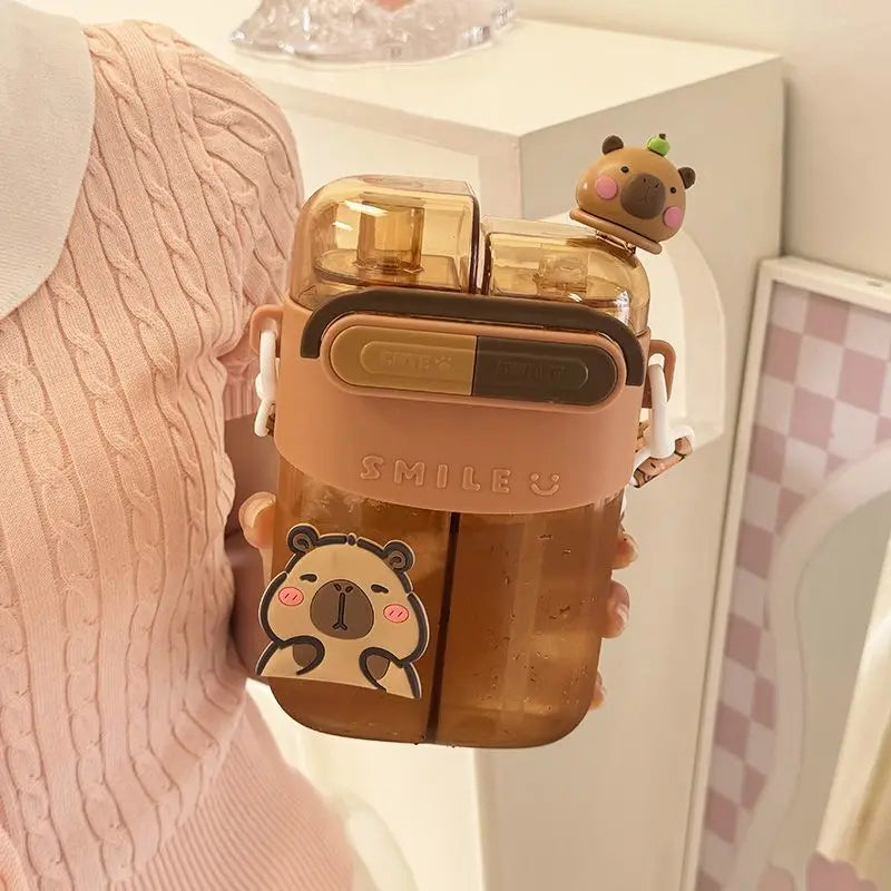 Cute Capybara Kids Water Bottle – Large Capacity Straw Cup A person in a pink cable-knit sweater holds a translucent amber water bottle with a peach-colored collar, cartoon bear graphics, and a small bear-shaped cap on the spout in a bright indoor setting.