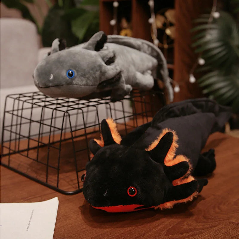 Cute Axolotl Plush Toy – Squishmallow Salamander Stuffed Animal Two plush axolotl toys displayed on a wooden table, with one gray plush resting on a black wire stand and one black plush with orange accents lying in front. The background shows a softly blurred indoor setting with plants and warm lighting.
