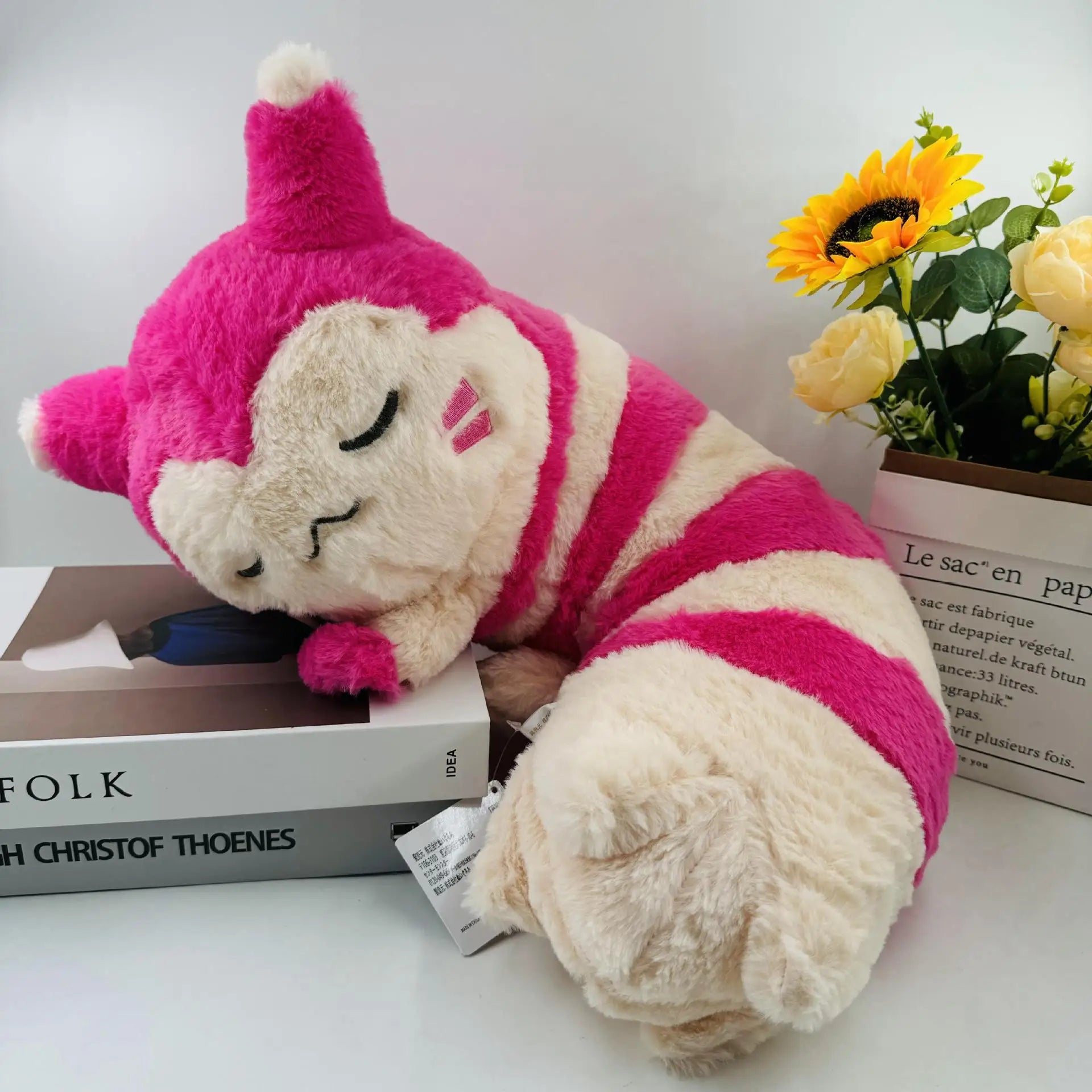 Pokemon Furret, Dratini & Snorlax Plush Neck Pillow A pink and cream striped plush toy character with a pointed hat, shown resting on a tabletop next to a book and a vase of yellow flowers.