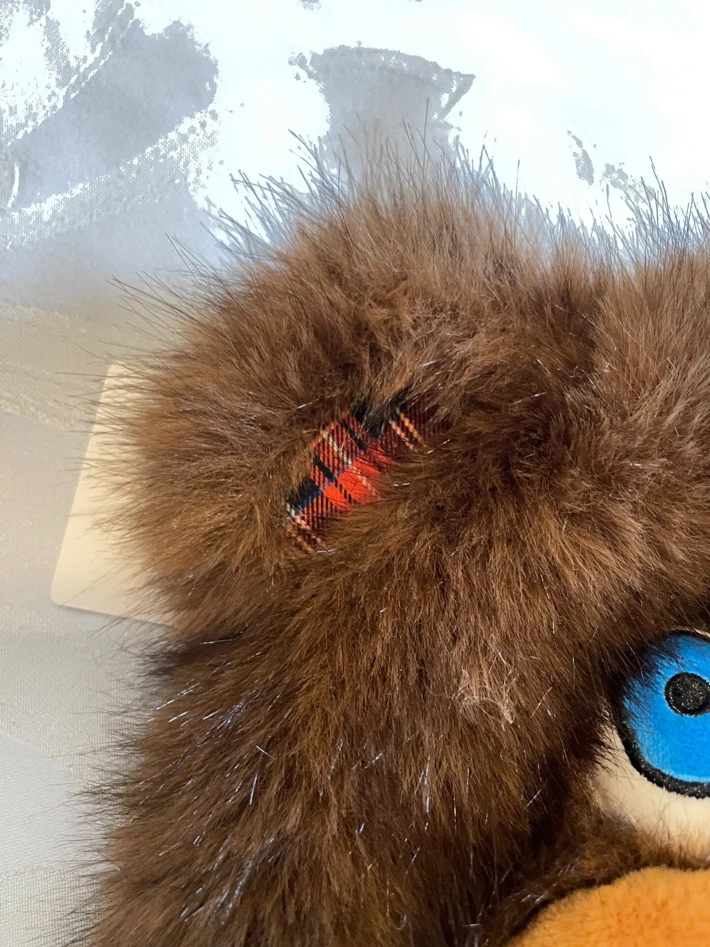 Hamikuma Bear Plush Hooded Halloween Trapper Hat Close-up of a brown faux fur accessory, likely a hat or plush toy head, featuring a small red and black plaid fabric accent and a blue eye detail.