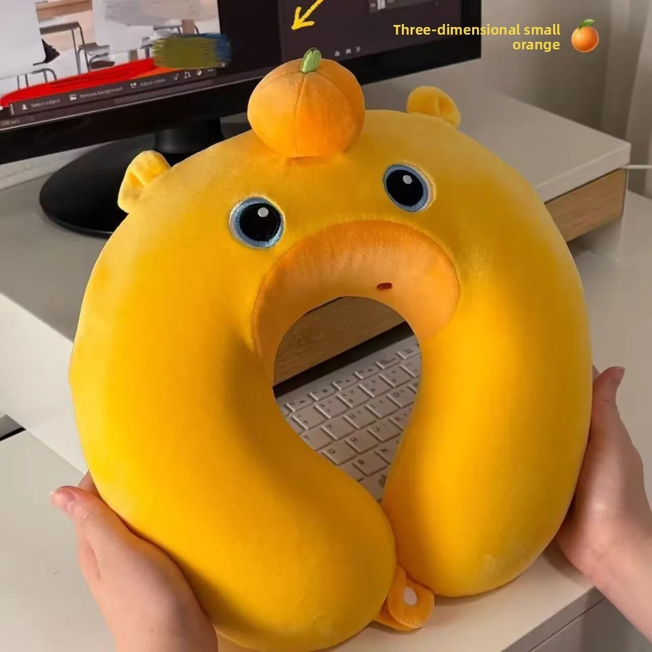 Cute Capybara U-Shaped Neck Pillow – Plush Travel Pillow Yellow neck pillow with cartoon face held by a person in front of a computer desk.