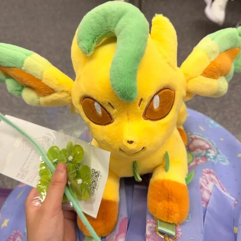 Pokemon Eevee Evolution Plush Crossbody Bag A yellow and green plush toy with large brown eyes, shown in a real-world setting with a hand holding a small green flower and a tag. The plush is attached to a purple patterned bag with green straps.