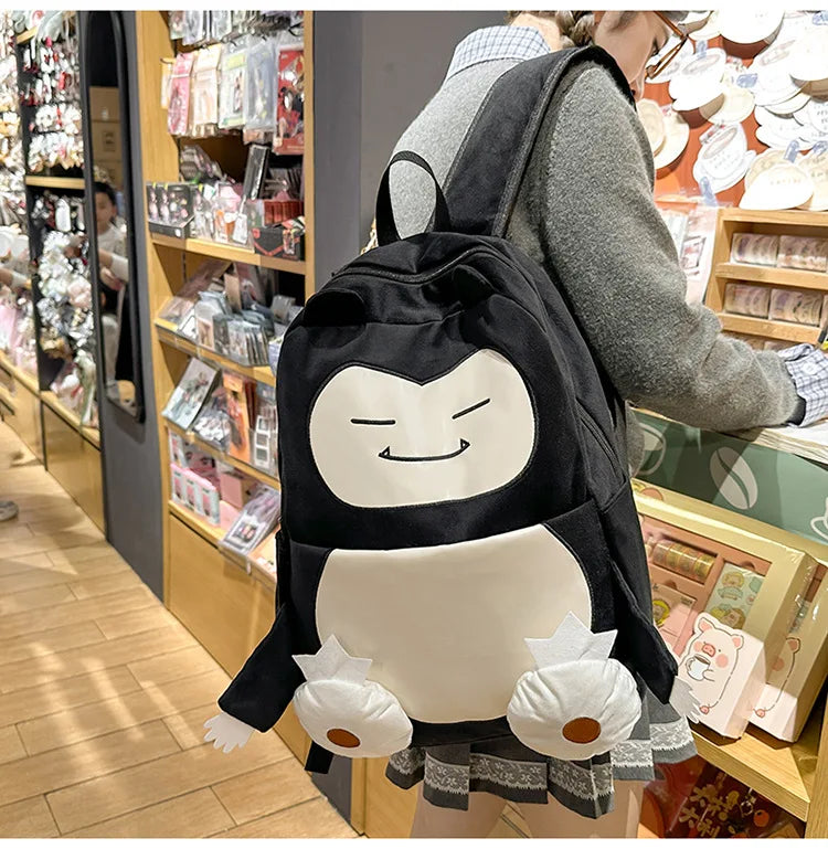 Snorlax Pokemon Backpack Large Capacity Casual Bag A person wearing a gray sweater carries a black and white penguin backpack in a retail store. The backpack features a cute cartoon penguin face and white flower-like paw pads.