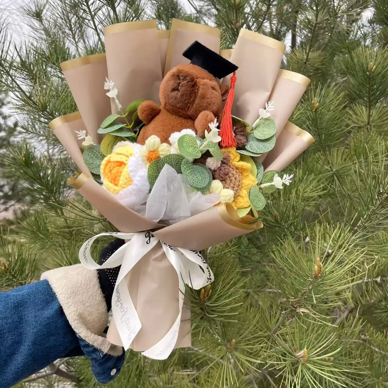 Capybara Plush with Crochet Flowers – Cute Flower Bouquet Gift Bouquet with a teddy bear wearing a graduation cap against a green foliage background