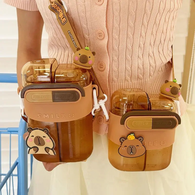 Cute Capybara Kids Water Bottle – Large Capacity Straw Cup A person wearing a pink cable-knit cardigan holds two translucent amber-brown double-walled water bottles with tan protective sleeves, cartoon bear decorations, and a matching shoulder strap.