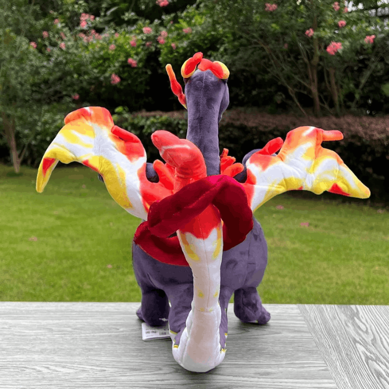Pokemon Charizard Plush Toy – Shiny & Gigantamax A plush toy dragon with purple body, red and orange flame details, and white wing-like flaps, photographed outdoors on a gray table with grass and bushes in the background.