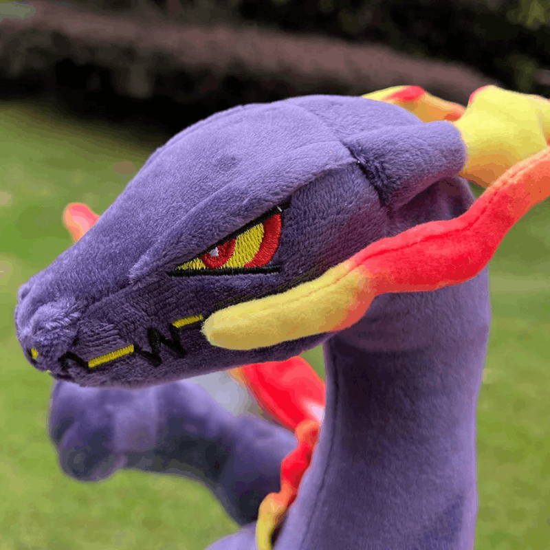Pokemon Charizard Plush Toy – Shiny & Gigantamax A close-up of a plush toy dragon in purple with yellow and red flame-like accents, photographed outdoors on a grassy background.