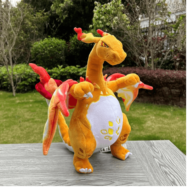 Pokemon Charizard Plush Toy – Shiny & Gigantamax A plush toy of Charizard, the orange dragon from Pokémon, sitting on a gray wooden table outdoors with a grassy garden and trees in the background.