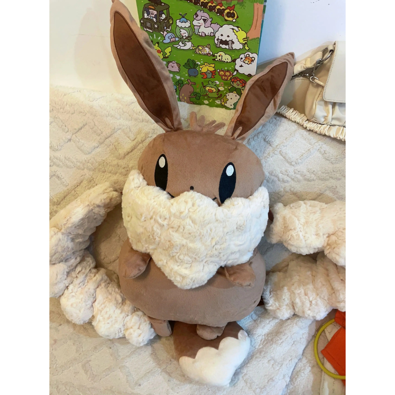 Eevee Plush Backpack – Kawaii Pokemon Cosplay Bag A brown plush toy with long upright ears, large black eyes, and a fluffy cream-colored scarf or wing detail, posed on a textured cream blanket with a colorful book in the background.