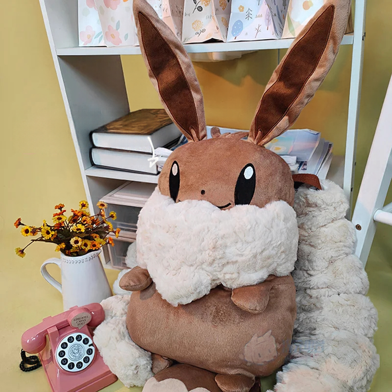 Eevee Plush Backpack – Kawaii Pokemon Cosplay Bag A plush toy shaped like a brown bunny with long ears, large black eyes, and a fluffy white scarf, styled on a shelf next to a pink rotary phone and small flower arrangement.