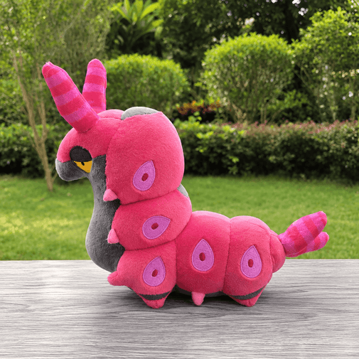 A pink and purple caterpillar plush toy resembling a Pokémon Larvestith, sitting on a gray wooden table with a blurred green garden background.