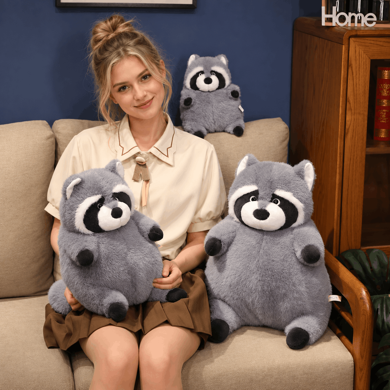 A woman sits on a beige sofa holding two gray raccoon plush toys, with a matching raccoon plush toy behind her on the couch. The setting includes a blue wall and a wooden cabinet in the background.