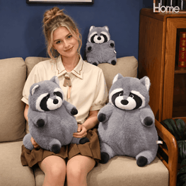A woman sits on a beige sofa holding two gray raccoon plush toys, with a matching raccoon plush toy behind her on the couch. The setting includes a blue wall and a wooden cabinet in the background.