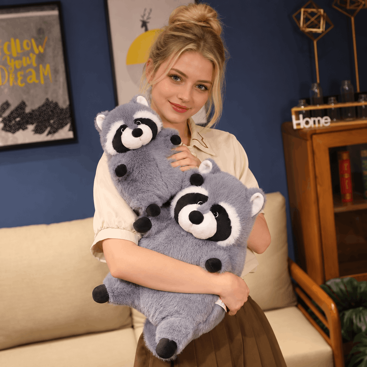A woman in a cream shirt and brown skirt holds a large gray plush raccoon toy with black and white facial features in a cozy living room setting.