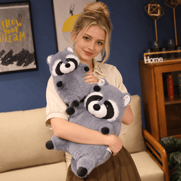 A woman in a cream shirt and brown skirt holds a large gray plush raccoon toy with black and white facial features in a cozy living room setting.