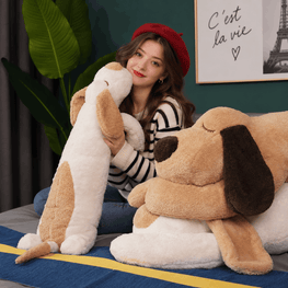 A woman wearing a red beret and striped sweater sits on a bed holding a large plush dog toy, with another oversized dog plush pillow beside her in a styled bedroom setting.