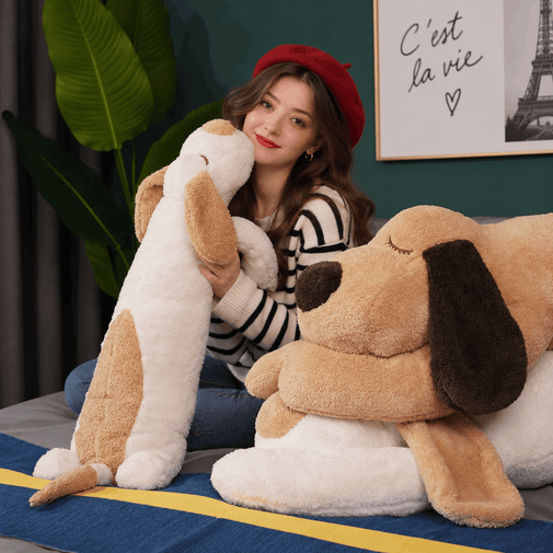 A woman wearing a red beret and striped sweater sits on a bed holding a large plush dog toy, with another oversized dog plush pillow beside her in a styled bedroom setting.