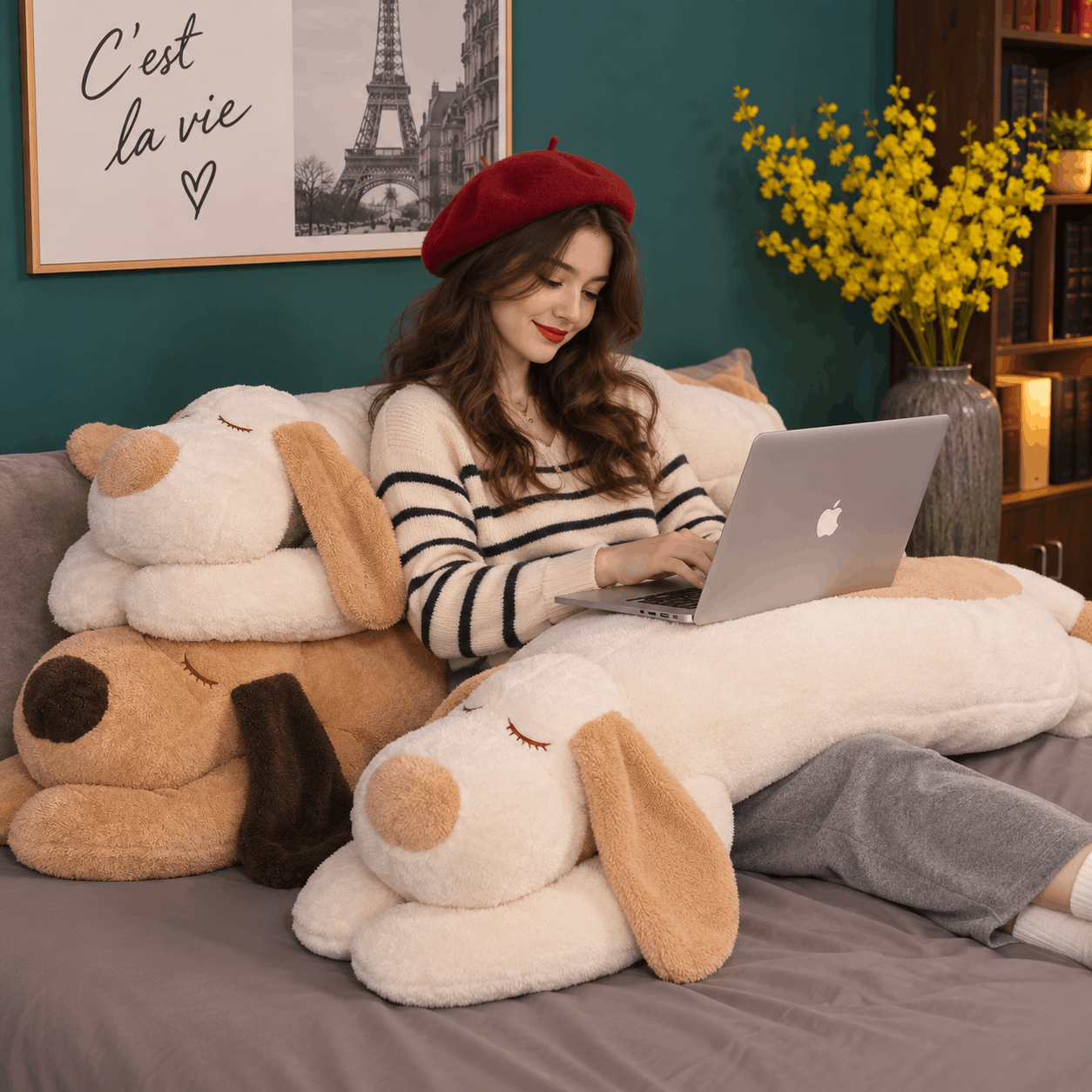 A woman sits on a bed surrounded by large plush dog toys, using a silver laptop. She wears a red beret, a black-and-white striped sweater, and gray pants.