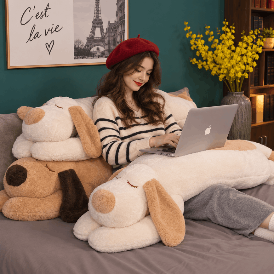 A woman sits on a bed surrounded by large plush dog toys, using a silver laptop. She wears a red beret, a black-and-white striped sweater, and gray pants.
