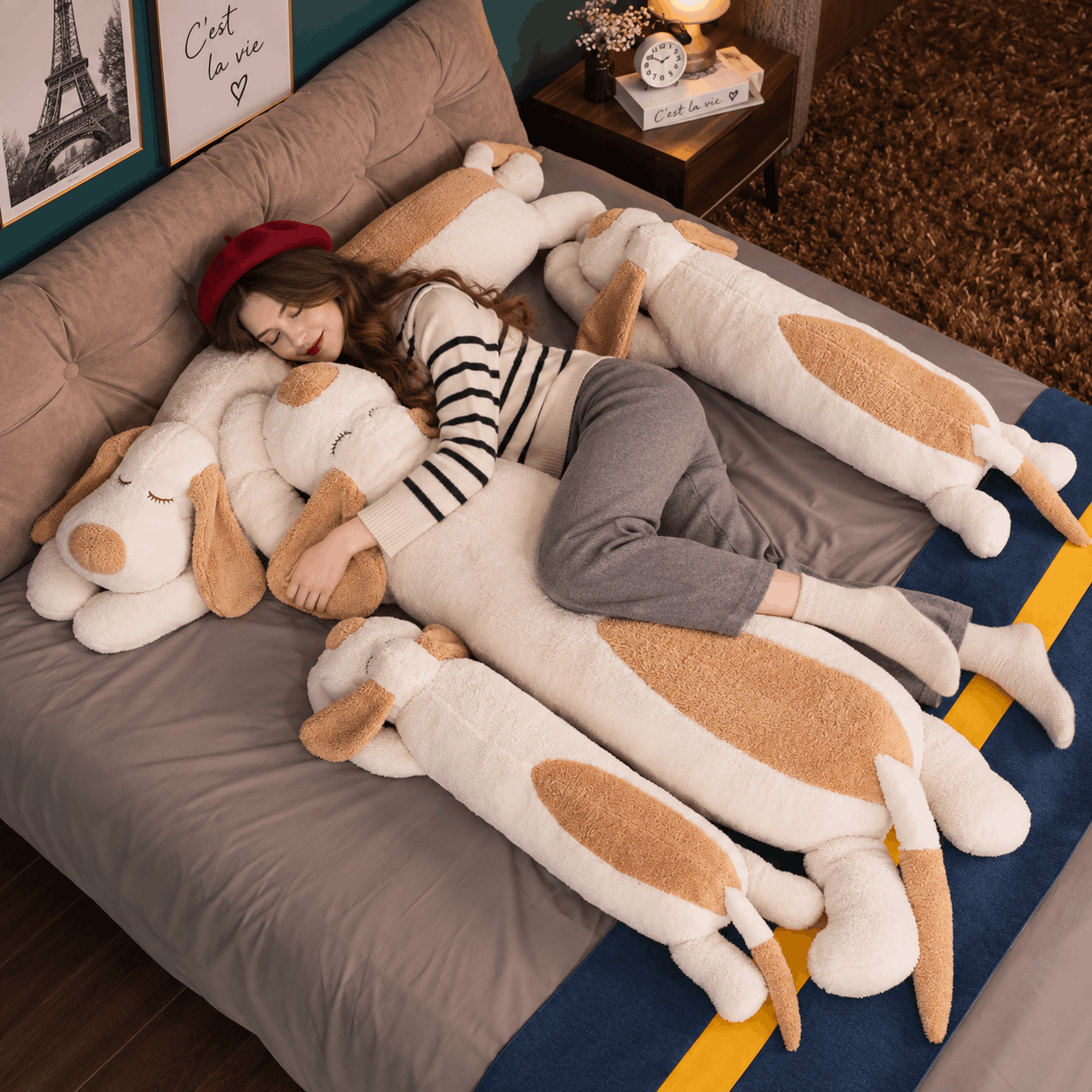 A woman lies on a bed surrounded by oversized plush dog-shaped pillows and a long dog-shaped body pillow in cream and tan, in a styled bedroom setting.