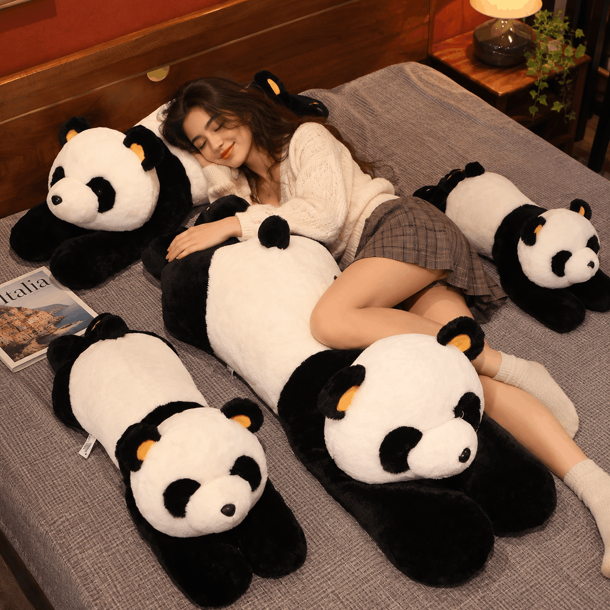 A woman lies on a bed surrounded by four large plush panda dolls in black and white, with small orange ear accents. The scene is indoors on a gray bedspread with wooden headboard and bedside lamp visible.