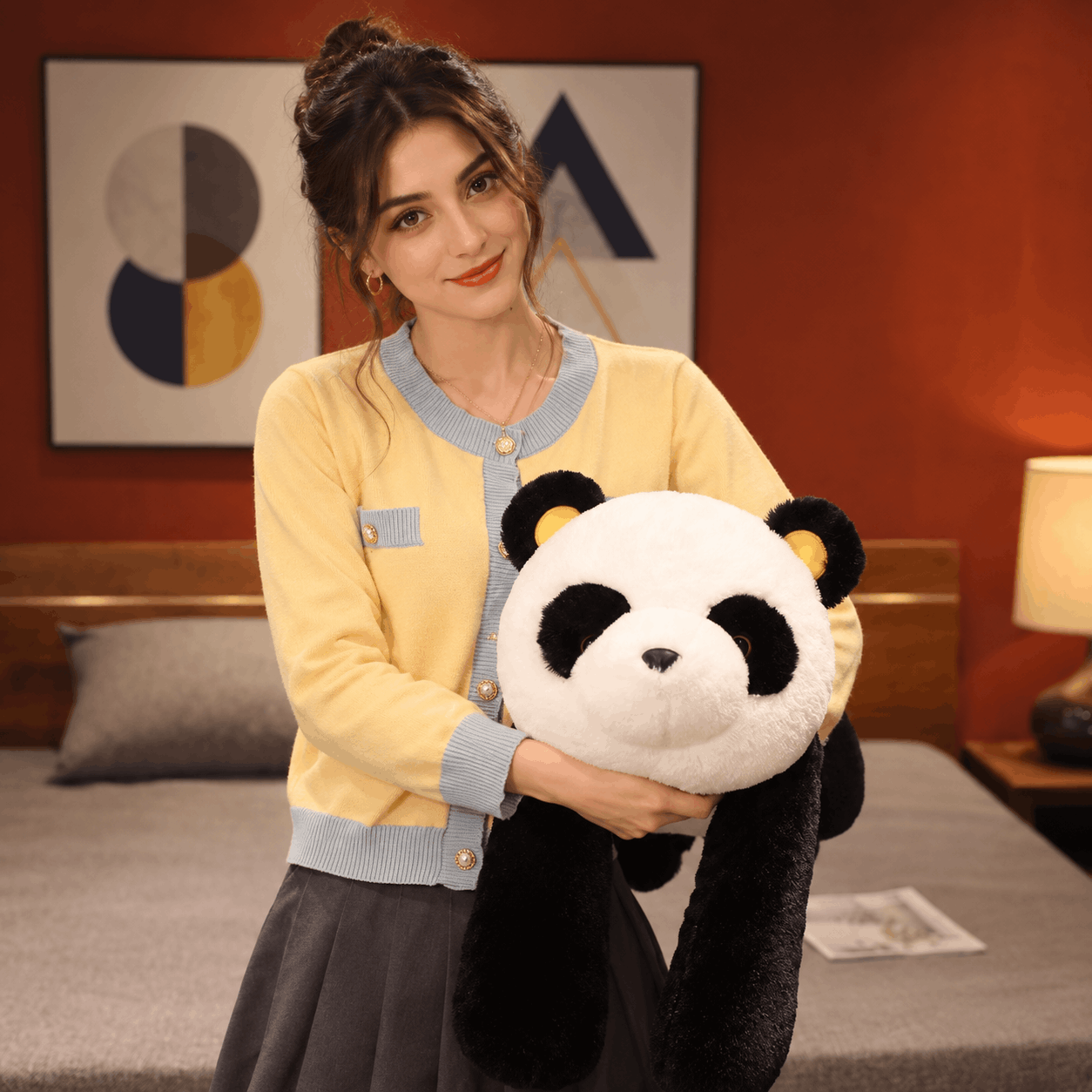 A woman in a yellow cardigan and gray skirt holds a large plush panda toy in a warmly lit bedroom setting.