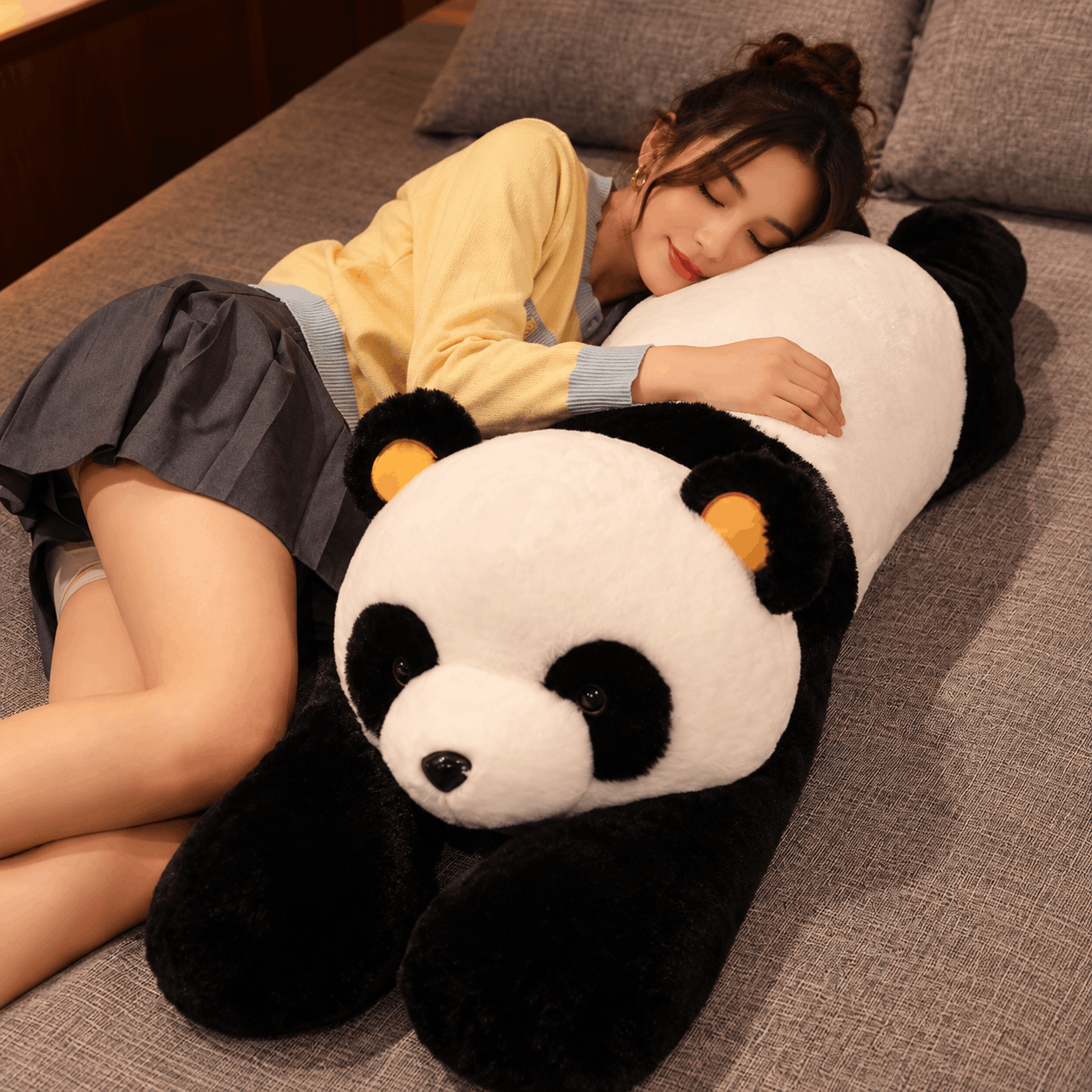 A woman lying on a gray sofa, resting her head on a large black-and-white panda plush toy with orange ear accents.