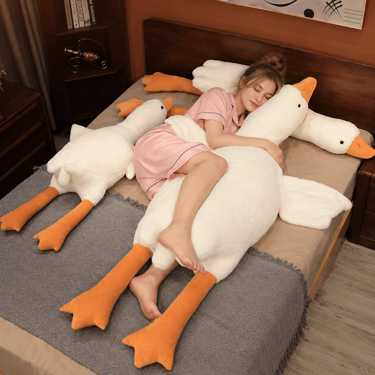 A woman in a pink pajama set lies on a bed with a large plush goose pillow, with two additional goose pillows on either side of the bed in a bedroom setting.