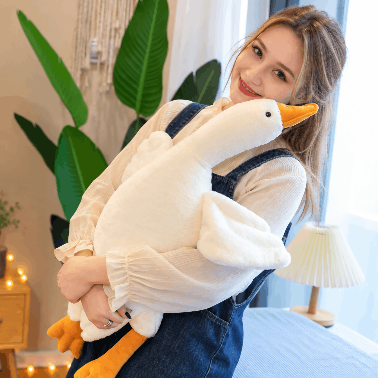 A woman in a bedroom setting holds a large plush white goose toy. She is wearing a white long-sleeve blouse and blue denim overalls, with a bed, lamp, and potted plant visible in the background.